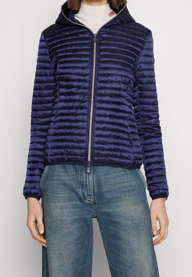 Femme portant une veste matelassée bleu marine zippée avec capuche, associée à un jean en denim bleu et un pull à col roulé blanc.