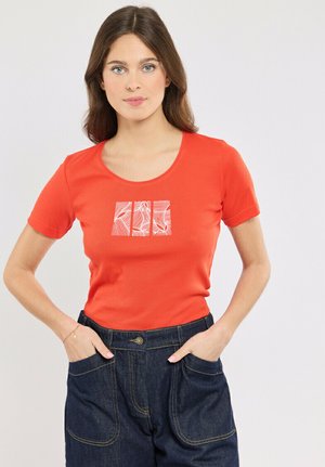 Jeune femme aux longs cheveux bruns portant un t-shirt rouge à manches courtes avec un motif abstrait en carrés blancs et un pantalon en denim bleu foncé, les mains dans les poches.