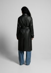 Trench coat de cuero negro con cinturilla, con cuello clásico y mangas largas, combinado con vaqueros de pierna ancha de color azul claro.