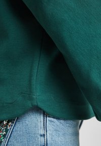 Grüner Baumwollpullover mit abgerundetem Saum, der Texturdetails zeigt. Über blauen Jeans mit sichtbaren Nähten getragen, aus denen ein florales Stoffmuster hervorschaut.