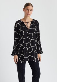 Zwarte blouse met lange mouwen en een wit geometrisch patroon, met een striksluiting bij de hals en een ontspannen fit. Gladde stof met omgeslagen mouwen.