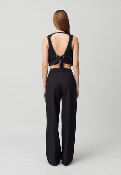 Top corto negro con un profundo corte en la espalda y un detalle de lazo en la cintura, combinado con pantalones negros de corte holgado. Textura suave, diseño minimalista.