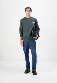 Homem a vestir um sweatshirt verde e umas calças de ganga azuis, segurando uma bolsa de couro preta em frente a um fundo branco liso.