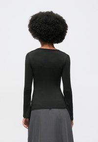 Vrouw met een afro-haarstijl, gekleed in een aansluitende zwarte lange mouwtop en een grijze rok, staand met haar rug naar de camera.