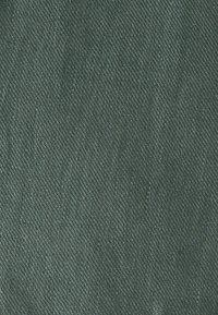 Nærbillede af grønt denimmateriale, der viser diagonal vævningsstruktur og detaljeret syningsmønster.