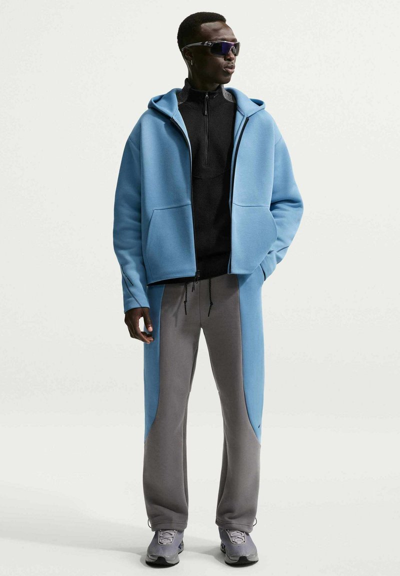 Mann trägt einen blauen Hoodie, ein schwarzes Zip-up-Shirt, graue Hosen mit blauen Seitenpaneelen, graue Turnschuhe und schwarze Sonnenbrille, steht vor weißem Hintergrund.