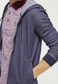 Felpa con zip color navy con accenti grigi, dotata di tasca frontale e cappuccio con cordoncino, indossata sopra una camicia bordeaux con colletto e motivo testurizzato.