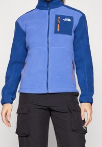 Niebieska polarowa kurtka z wysokim kołnierzem, kieszenią na zamek błyskawiczny na piersi oraz kontrastującymi niebieskimi rękawami. Zawiera pomarańczowe akcenty na zamkach.