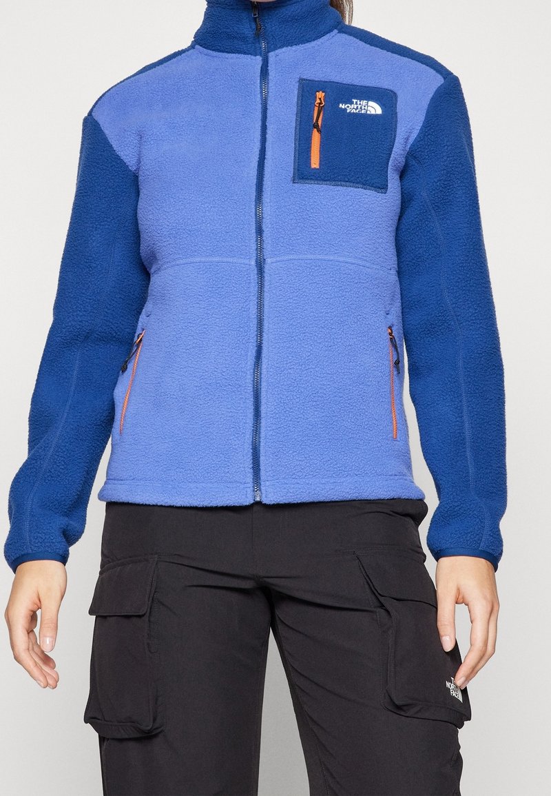 Niebieska polarowa kurtka z wysokim kołnierzem, kieszenią na zamek błyskawiczny na piersi oraz kontrastującymi niebieskimi rękawami. Zawiera pomarańczowe akcenty na zamkach.