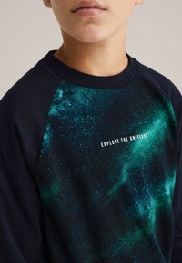 Felpa blu navy con un motivo galattico in tonalità di verde e blu, con il testo "ESPLORA L'UNIVERSO" al centro.