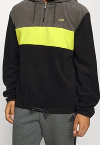 Sort og grå lynlås hoodie med neon gul horisontal stribe, med snoretræk i hætten og logo på brystet. Blød tekstur.