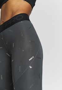 Legginsy Nike Pro w ciemnoszarym kolorze z czarnym paskiem w talii. Charakteryzują się gładką teksturą oraz powtarzającym się odblaskowym wzorem z logo.