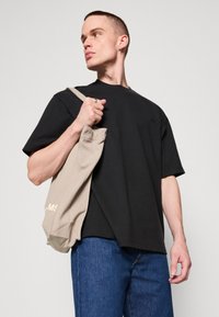 Tricou negru din bumbac cu guler rotund, purtat cu blugi albaștri. Ținând o geantă din pânză bej, cu un branding minim.