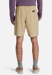 Beige cargo šortky s elastickým pasem, bočními kapsami a malou nášivkou s logem. Nosit s šedými ponožkami a světlejší obuví.