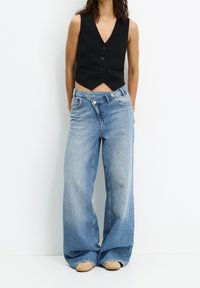 Gilet noir sans manches avec un décolleté en V et des boutons à l'avant, associé à un jean large léger bleu clair et des espadrilles beiges.