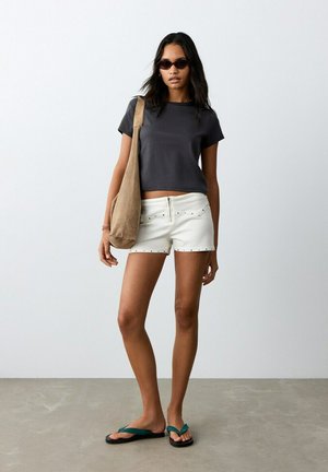 Jeune femme portant des lunettes de soleil, un t-shirt noir court, un short blanc clouté, des tongs turquoise, et portant un grand sac à épaule beige.