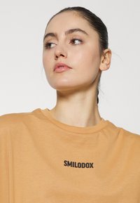 Camiseta laranja clara com decote redondo, feita de material suave, apresentando um logótipo impresso em preto "SMILODOX" no peito.