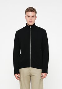 Nuori mies, jolla on päällään musta vetoketjullinen villapaita ja vaaleat cargo-housut, seisoo yksinkertaista valkoista taustaa vasten.