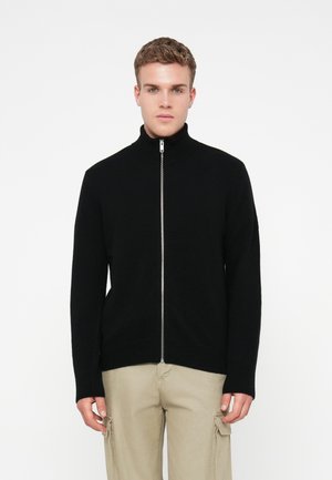 Jonge man in een zwarte ritsvest en beige cargobroek, staand tegen een effen witte achtergrond.