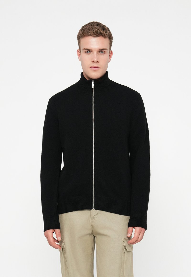 Noor mees mustas lukuga svetrisse ja beežides cargo-pükstes, seisev ühevärvilise valge tausta ees.