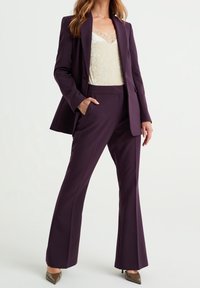 Completo tailored viola composto da un blazer aderente e pantaloni a zampa d'elefante. Sotto, una canotta di crema chiara con pizzo aggiunge texture.