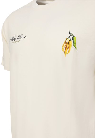 Cremefarbenes T-Shirt mit gestickten gelben und grünen Chilischoten auf der Brust und schwarzem, kursivem Text auf der gegenüberliegenden Seite.