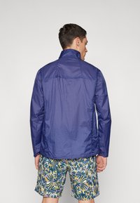 Giacca leggera blu navy con colletto alto e maniche lunghe, realizzata in materiale liscio e resistente all'acqua. Indossata sopra pantaloni corti a fantasia.