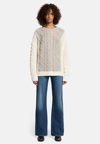 Pullover in maglia grossa a treccia di colore crema e beige, con un design strutturato; abbinato a jeans in denim blu a zampa d'elefante e scarpe nere.