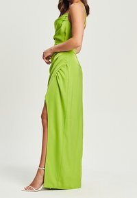 Kobieta ubrana w limonkowo zieloną satynową sukienkę bez rękawów z bocznym rozcięciem, w połączeniu z białymi sandałami na obcasie, stojąca na neutralnym tle.