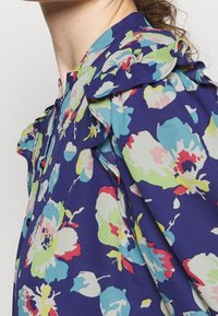 Květinová blůza v námořnické modři s živými, multicolor květinovými vzory. Obsahuje volány na ramenou a knoflíkový uzávěr vpředu.