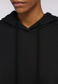 Svart hoodie i mjukt tyg med V-formad halsringning, dragsnören och en broderad logotyp på bröstet. Slät textur och minimalistisk design.