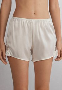 Pantalones cortos de satén color crema con un ajuste relajado, que presentan detalles de encaje y malla en los lados. Cintura elástica para mayor comodidad.
