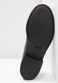 Semelle de chaussure noire avec texture antidérapante, logos "Camper" et "X Ultra Light" visibles sur le talon, positionnée contre un mur et un sol blancs.