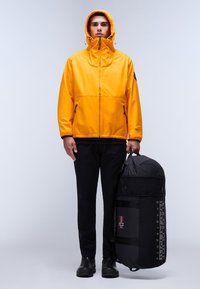 Žlutá nepromokavá bunda s vysokým límcem, černými zipy a podšívkovou kapucí. Černá sportovní taška s logem a dvoubarevným designem, držena v ruce.