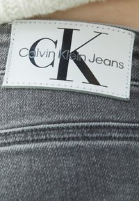 Grå denimtyg med en vit läderetikett som visar "Calvin Klein Jeans" och en framträdande "CK"-logotyp. Sömmar synliga längs kanterna.