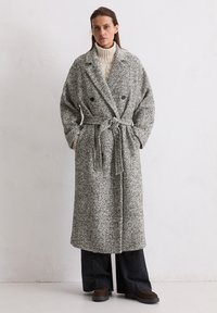 Grijs en zwart gespikkelde lange jas met een dubbelgestreept ontwerp, brede mouwen, tailleband en textuurstof, gedragen over een crèmekleurige trui.
