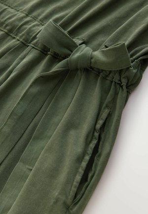 Rochie verde dintr-un material moale cu talie legată, având pliuri și buzunare laterale. Textură netedă și siluetă relaxată.