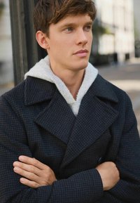 Young man with folded arms wearing a dark textured coat and light gray hoodie, looking thoughtfully to the side outdoors.