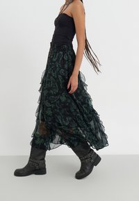Zwarte strapless top gecombineerd met een lange, doorschijnende zwarte rok met groene bloemenpatronen, afgewerkt met kniehoge zwarte laarzen met gespen.