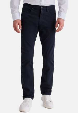 Mann trägt dunkelblaue, gerade geschnittene Hose, weiße Sneakers und ein weißes Hemd mit kleinen Punkten.