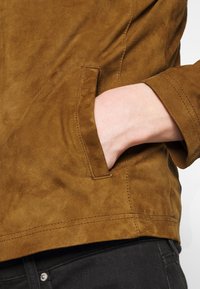 Brun mockaskinnjacka med sidofickor och avslappnad passform. Texturen verkar mjuk, med synlig sömnad längs nederkant och ärmar.