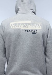 Sweatshirt à capuche gris avec un design imprimé au dos, affichant "NAPAPIJRI" et "EXP.87" dans des couleurs contrastées et des éléments graphiques.