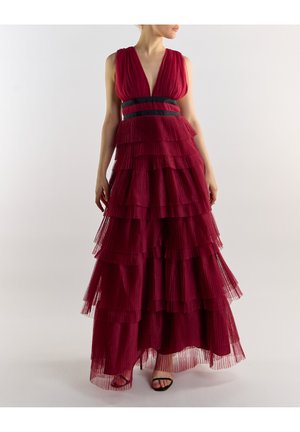 Mujer con un vestido sin mangas de tul en capas de un rojo profundo, con escote pronunciado y cintura con rayas negras, posando frente a un fondo sencillo.