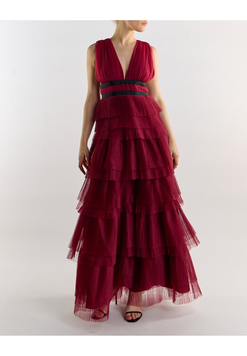 Kvinde iført en ærmeløs dybrød lagdelt tylkjole med dyb udskæring og sortstribet taljebånd, stående mod en ensfarvet baggrund.