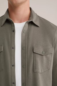 Primer plano de una persona que lleva una camisa de botón de oliva claro sobre una camiseta de cuello redondo blanca, mostrando detalles del cuello y del bolsillo en el pecho.