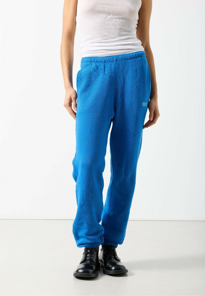 Osoba ubrana w jaskrawo niebieskie spodnie dresowe ze ściągaczami, czarne sznurowane buty oraz biały bezrękawnik na jednolitym białym tle.