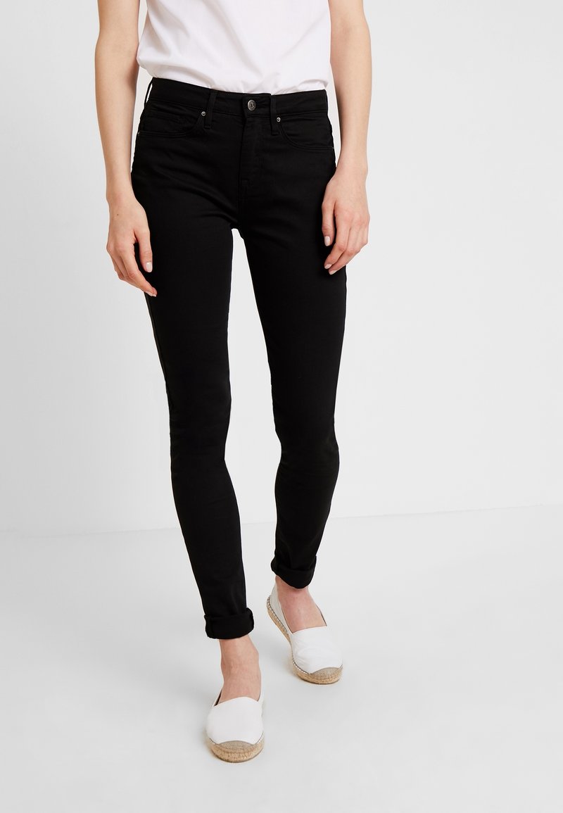 Jean skinny noir à texture lisse, taille mi-haute, poches avant et revers retroussés, associé à des espadrilles blanches.