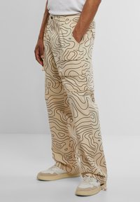 Beige cargo pants with a wavy black line pattern, featuring side pockets and a relaxed fit, paired with white and beige sneakers.
