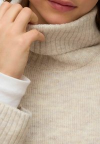 Bež rebrast pulover z visokim ovratnikom, izdelan iz mehkega pletenega materiala, z dolgimi rokavi, nošen čez belo obleko.