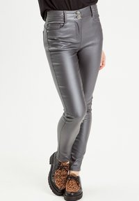 Pantalons skinny métalliques gris taille haute avec des détails cousus, deux boutons à l'avant et une texture lisse. Portés avec des chaussures à imprimé animalier.